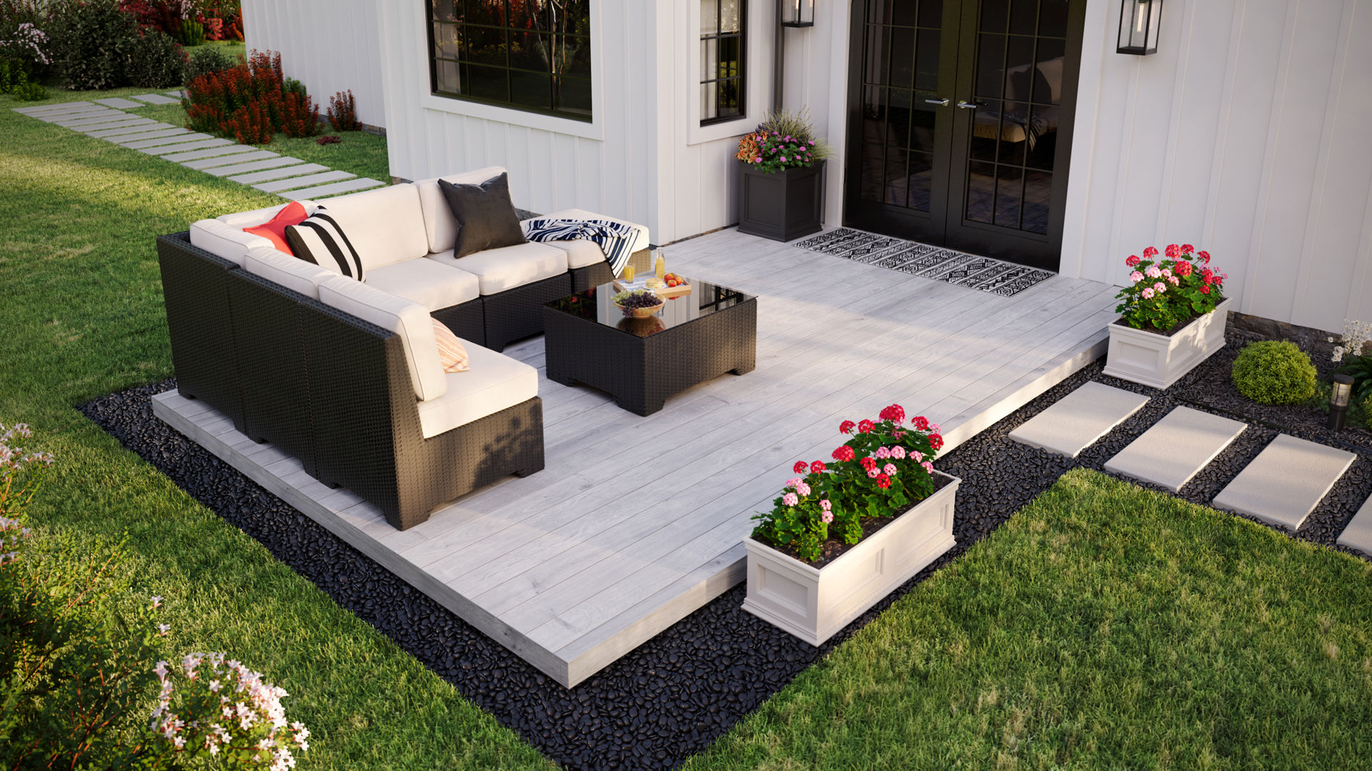 From a slightly raised angle, the image shows a deck attached to a white house. There is furniture in the left corner and a flower pot on the right. The deck sits atop gravel surrounded by grass.