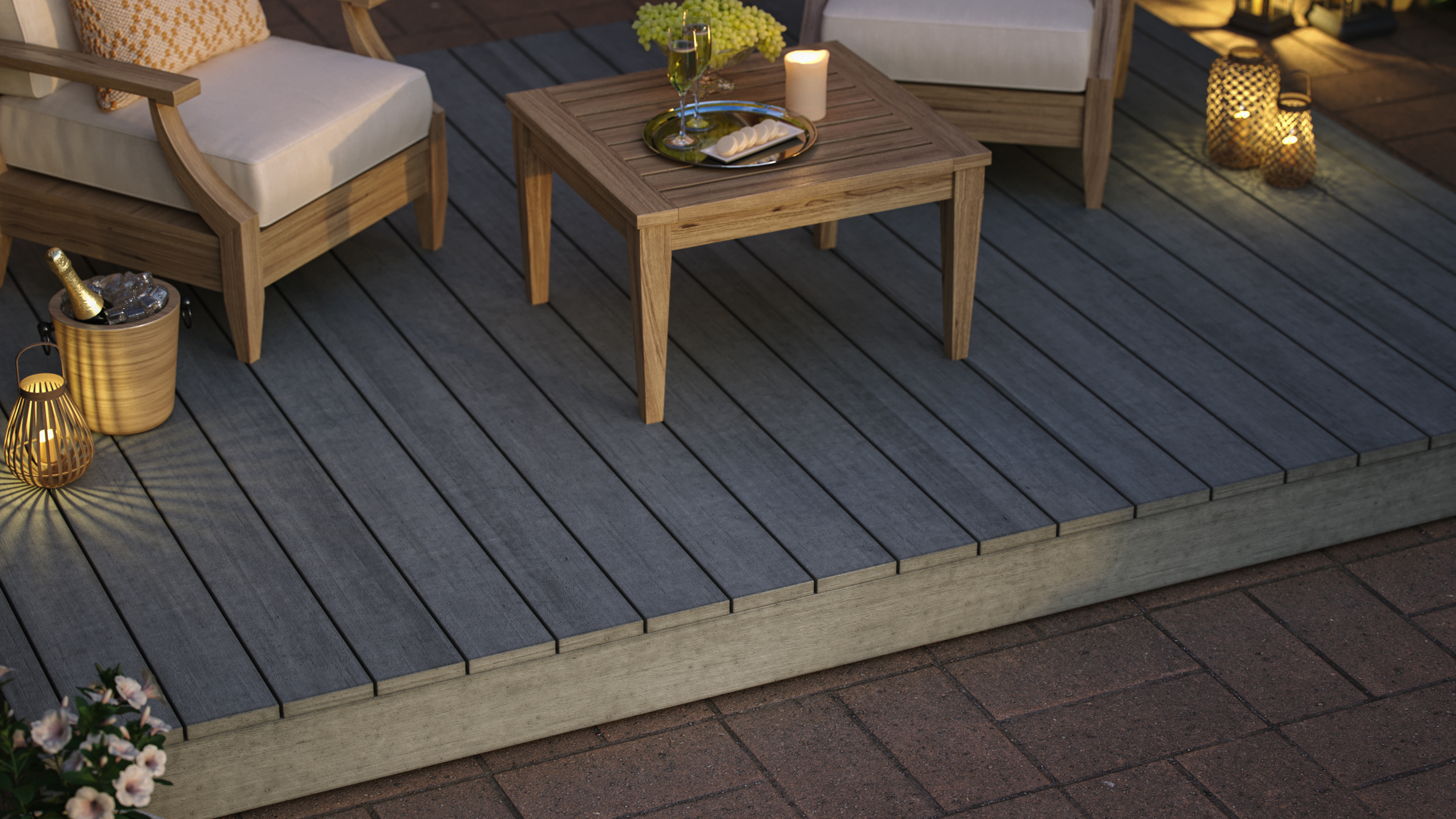 The image shows a deck with two chairs and a table. There is a tray, candle and centerpiece on the table. There are lanterns, champagne and flowers off to the sides, on the ground. The deck is on top of brick floors. 